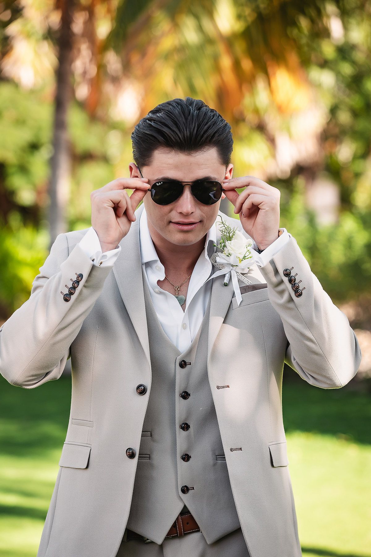 Groom in light gray suit and sunglasses poses playfully in a tropical garden setting.