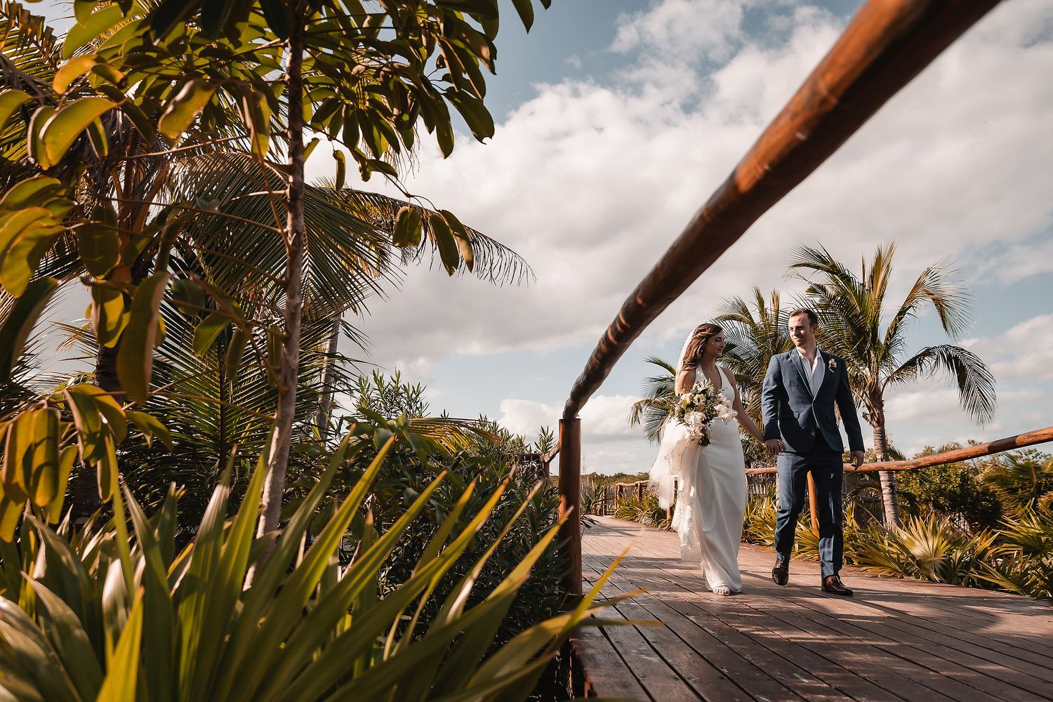Palm trees and lush foliage frame a wooden walkway under bright blue skies.