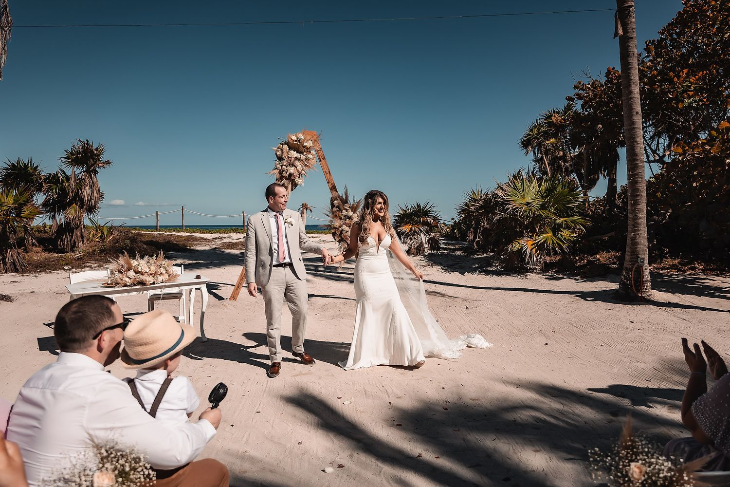 Guests celebrating at a beachside wedding with palm trees and clear blue skies.