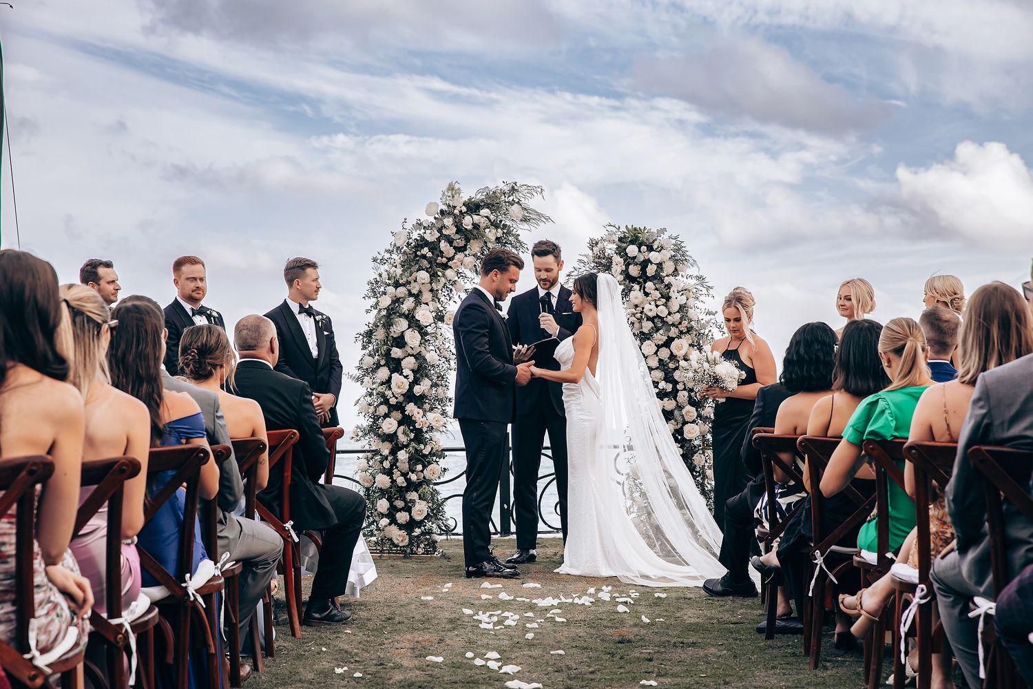 Wedding ceremony under a white floral arch with guests seated on both sides.
