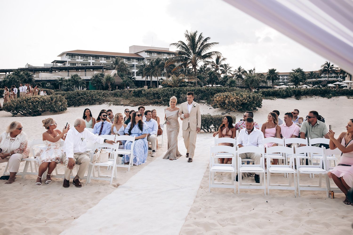 Wedding guests seated in white chairs along beachfront with palm trees in background.