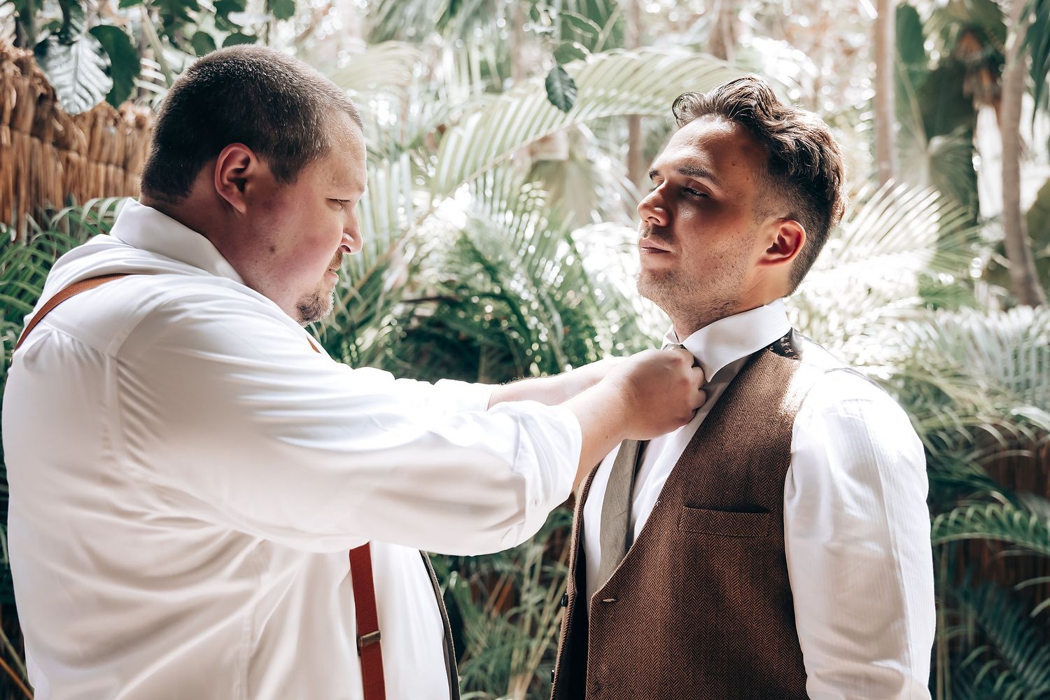 Two people in formal attire stand in a tropical garden setting while adjusting a tie.