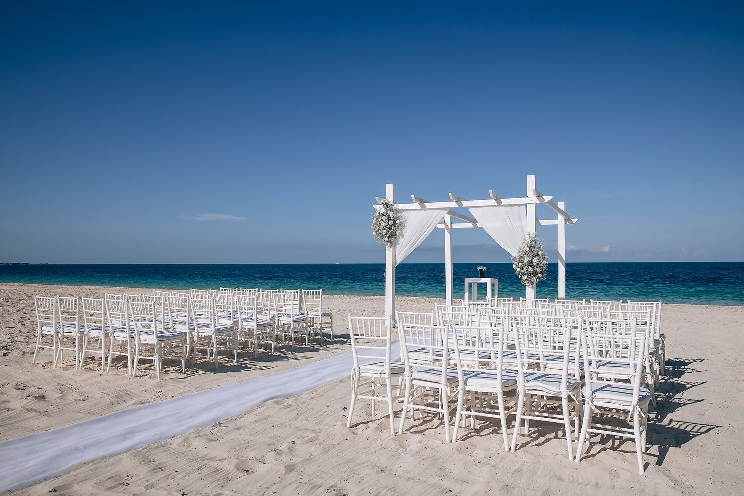 White wooden chairs and arch setup for a beach wedding ceremony with turquoise ocean backdrop.