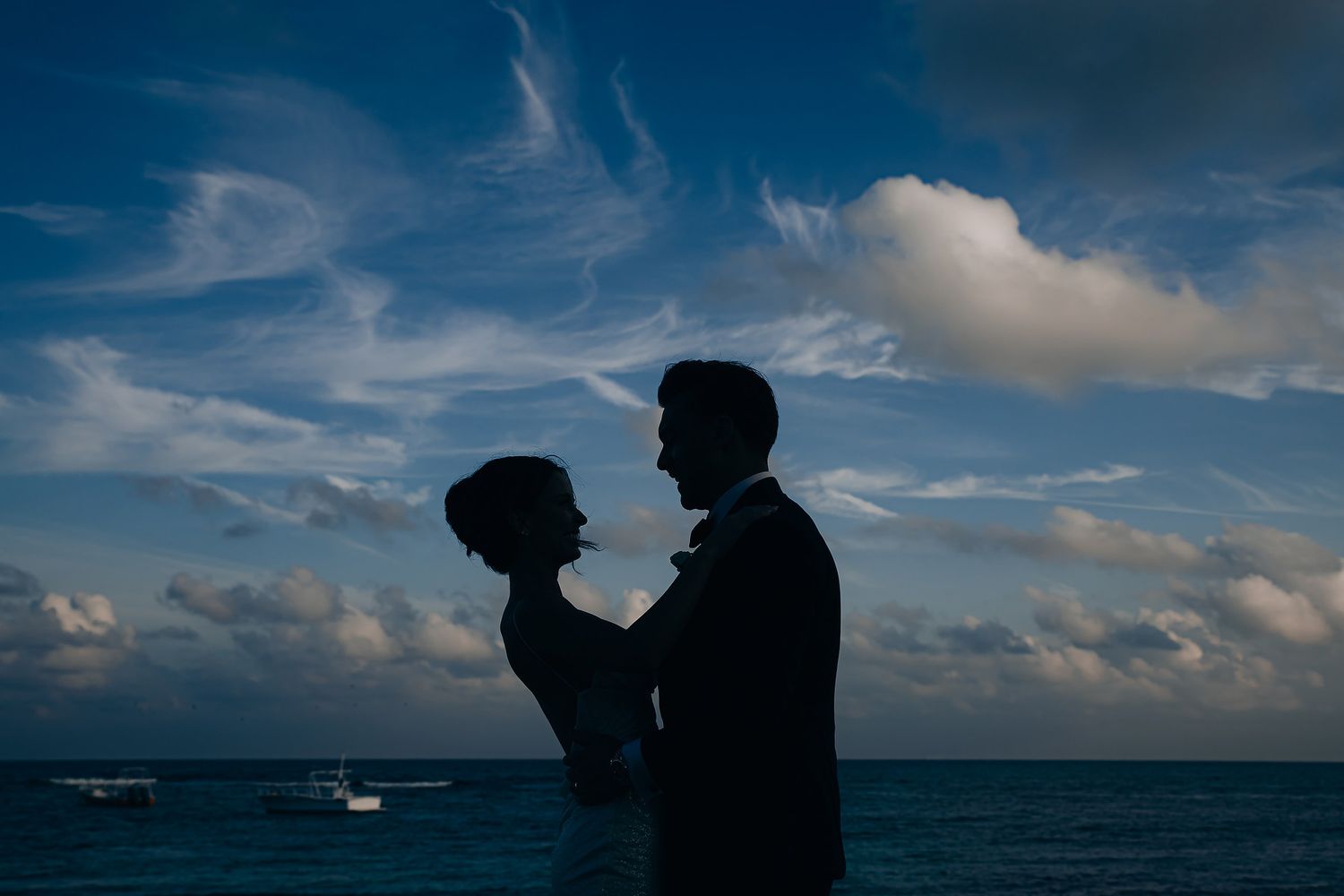 Silhouetted couple embracing against a dramatic blue sky with ocean and boats in the background.