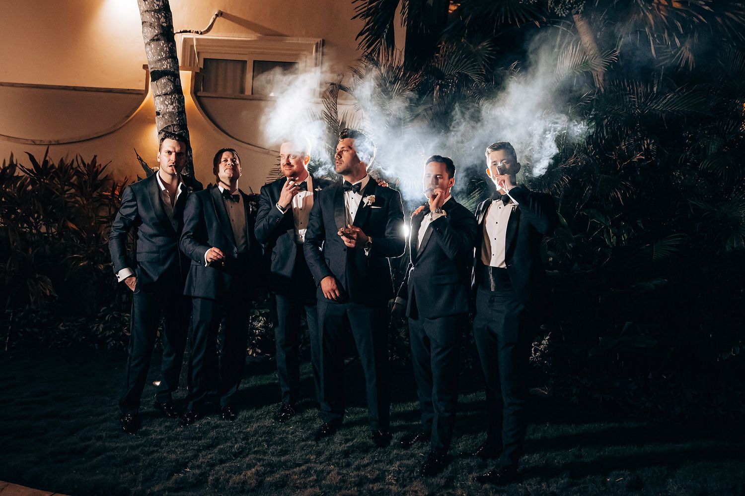 A group in formal attire stands together outdoors at dusk with dramatic lighting and trees in the background.