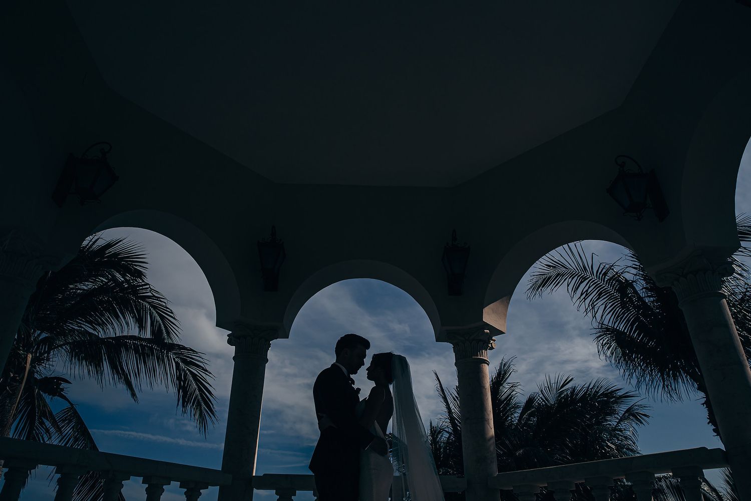 Silhouetted couple embracing under arched walkway with palm trees and moody blue sky at dusk.