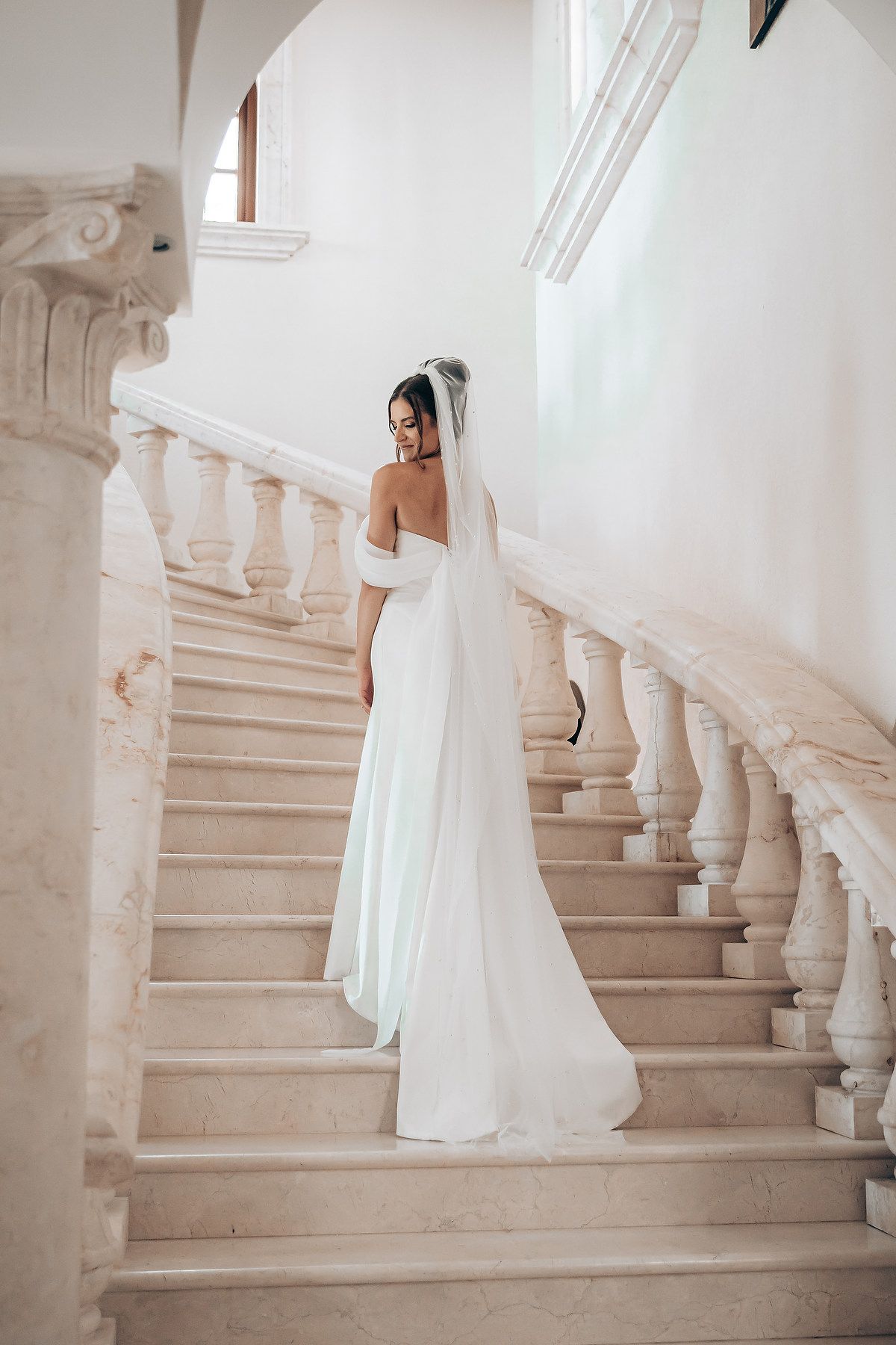 Elegant bride in flowing white dress and veil poses on grand marble staircase in bright interior setting.