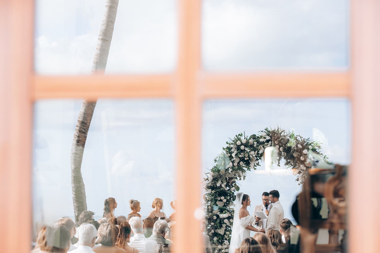 Outdoor wedding ceremony setup with pink-tinted columns and tropical landscaping against a cloudy sky.