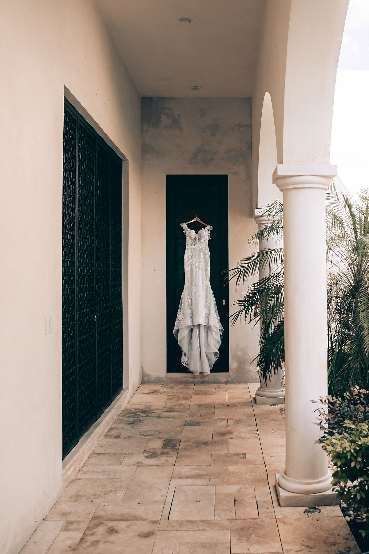 A white wedding dress hangs elegantly in a Mediterranean-style hallway with stone floors and white columns.