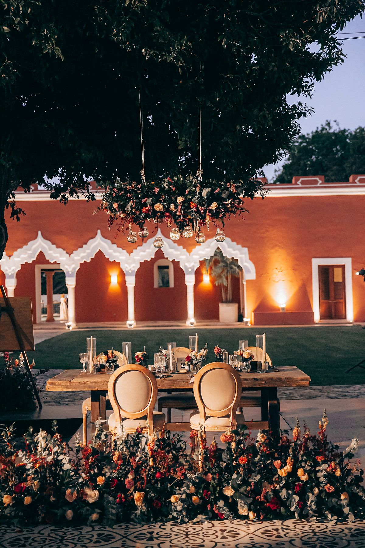 Rustic outdoor wedding reception setup with illuminated chairs and floral decorations against a terracotta building.