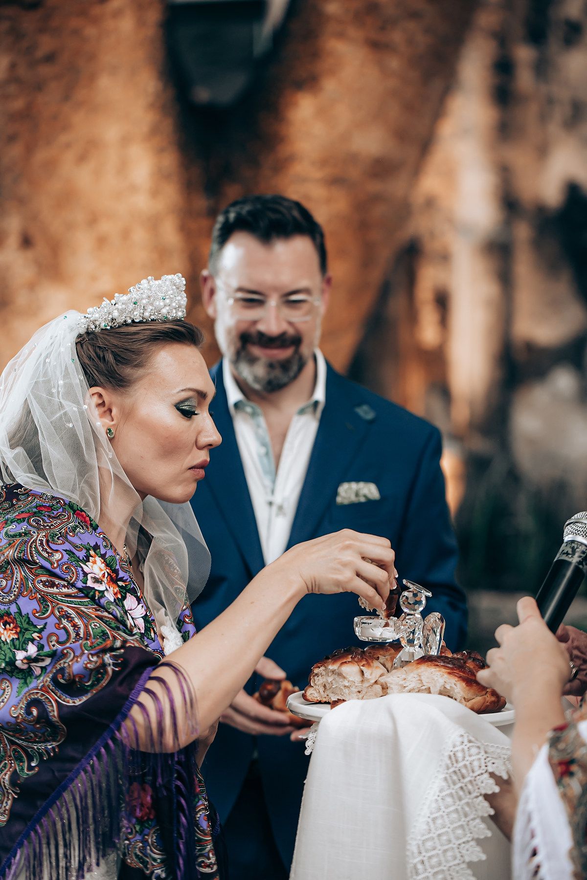 Traditional wedding celebration with couple sharing bread during ceremony against rustic stone wall backdrop.