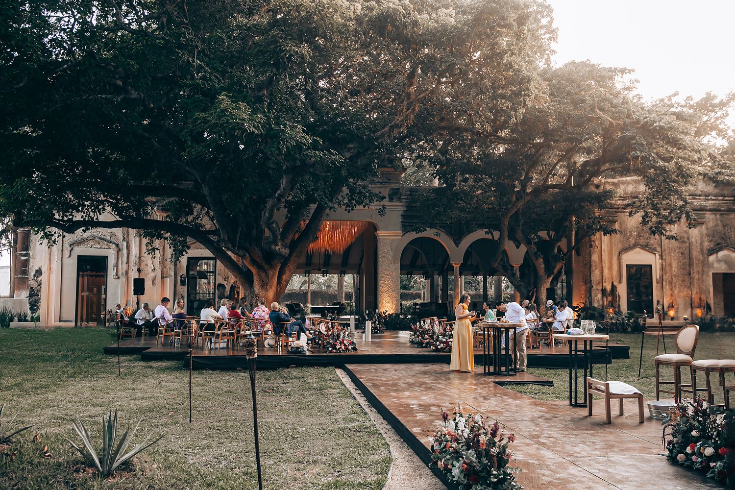 Scenic outdoor wedding venue setup with white chairs and floral arrangements under trees at sunset.