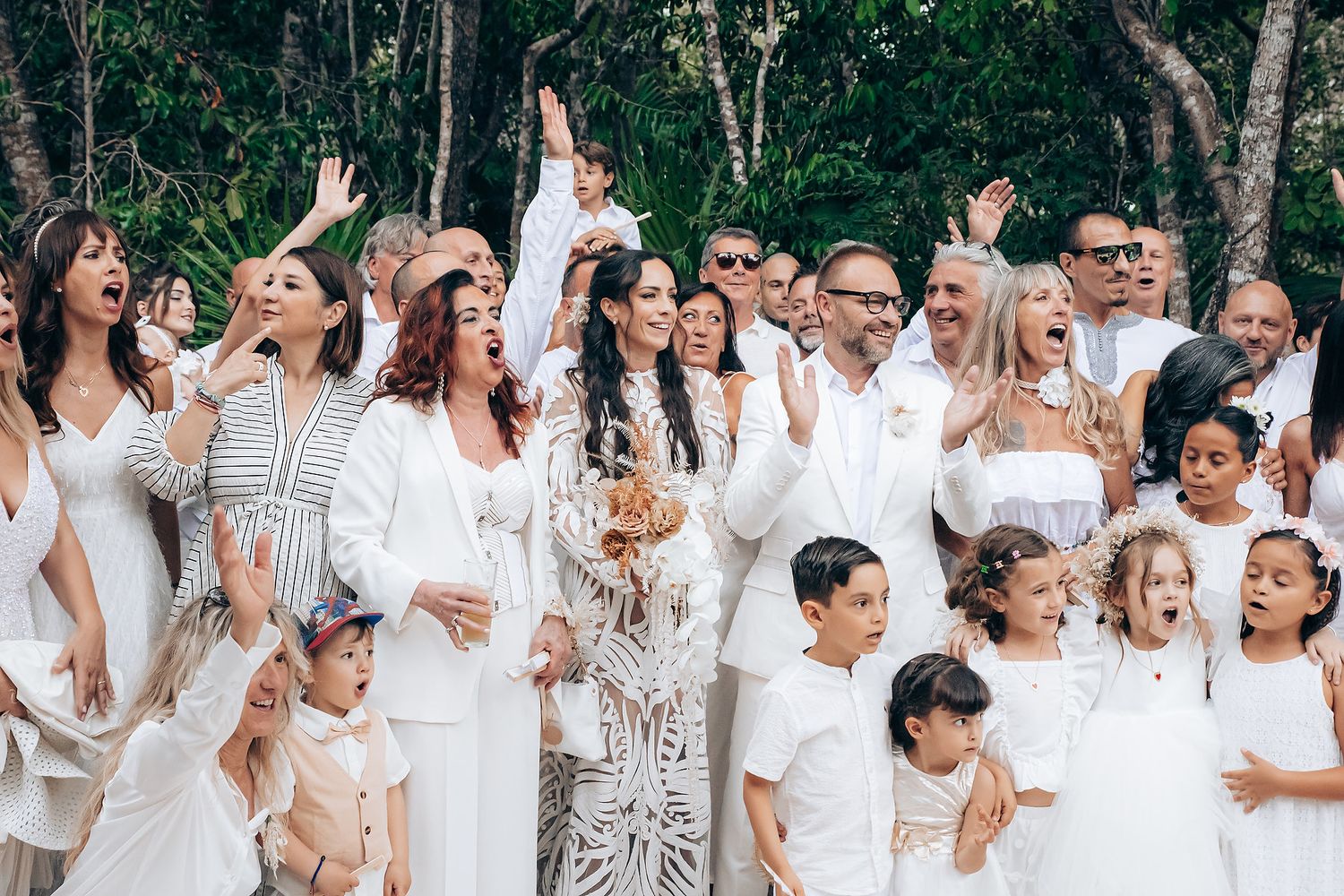 A large group wearing all-white outfits poses together for a celebratory outdoor photo under trees.