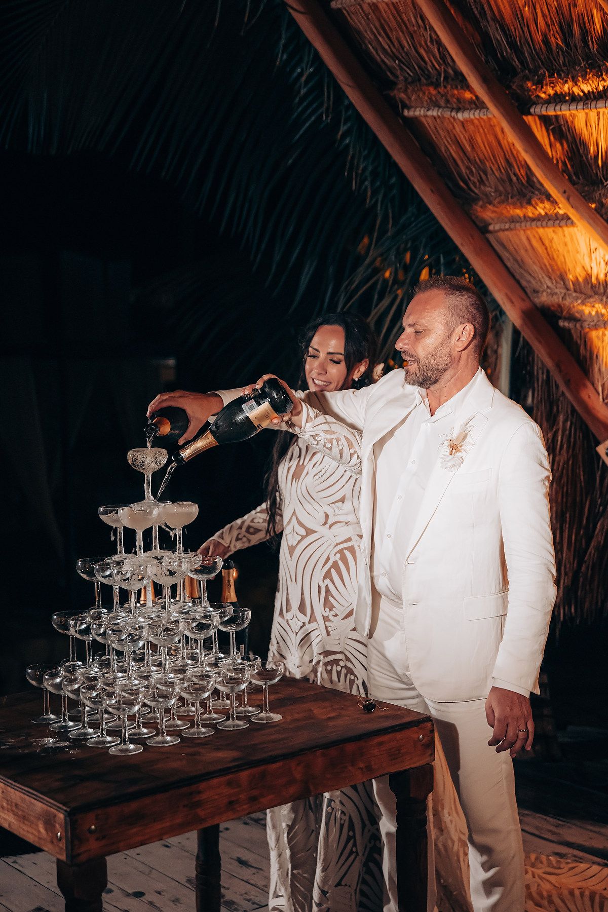 Two people in white attire pour champagne into a tower of elegant crystal glasses at a dimly lit event.
