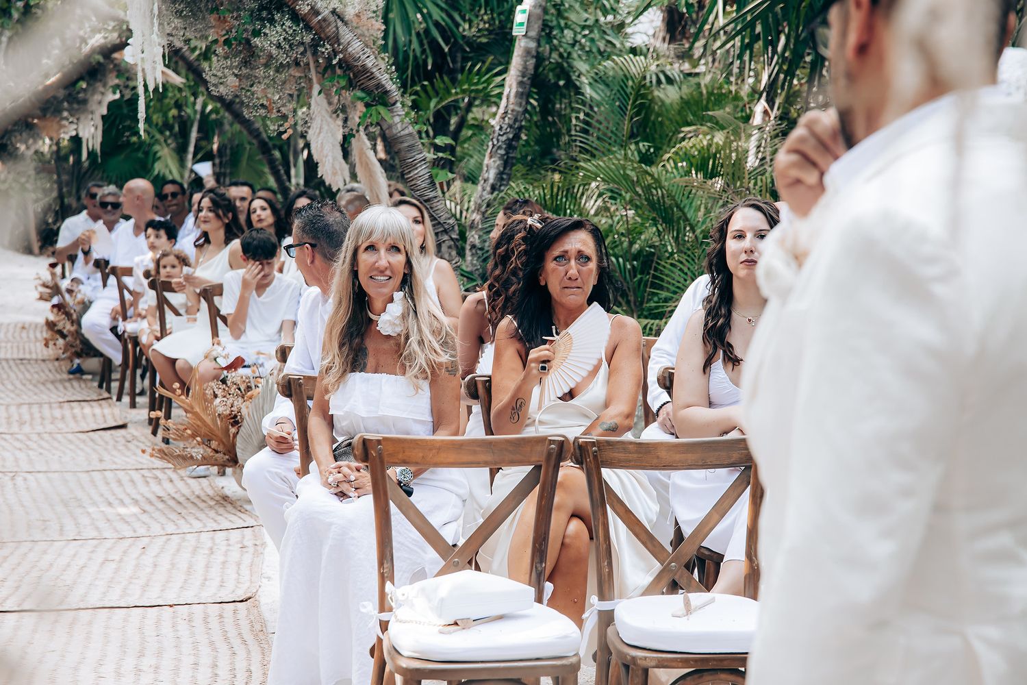 Wedding guests in white dresses gather outdoors at a tropical destination wedding ceremony with palm trees.
