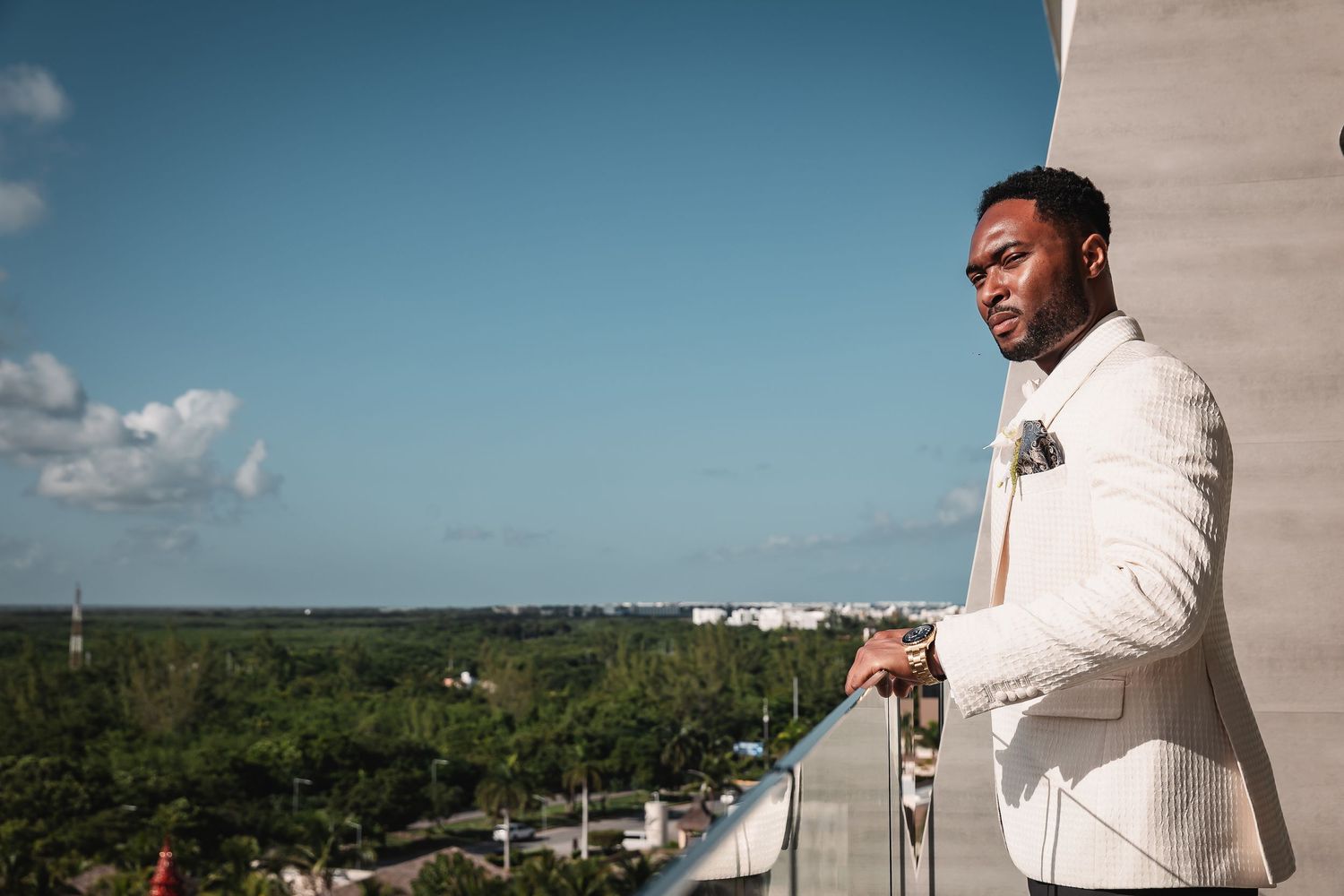 A person in a white tuxedo jacket stands on a balcony overlooking a scenic tropical landscape.