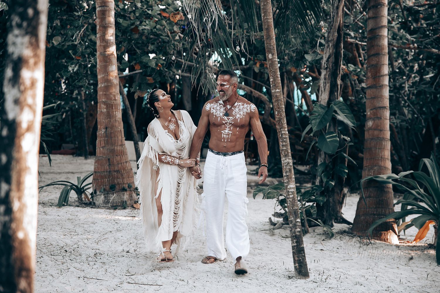 A couple strolls hand in hand along a sandy path through palm trees.