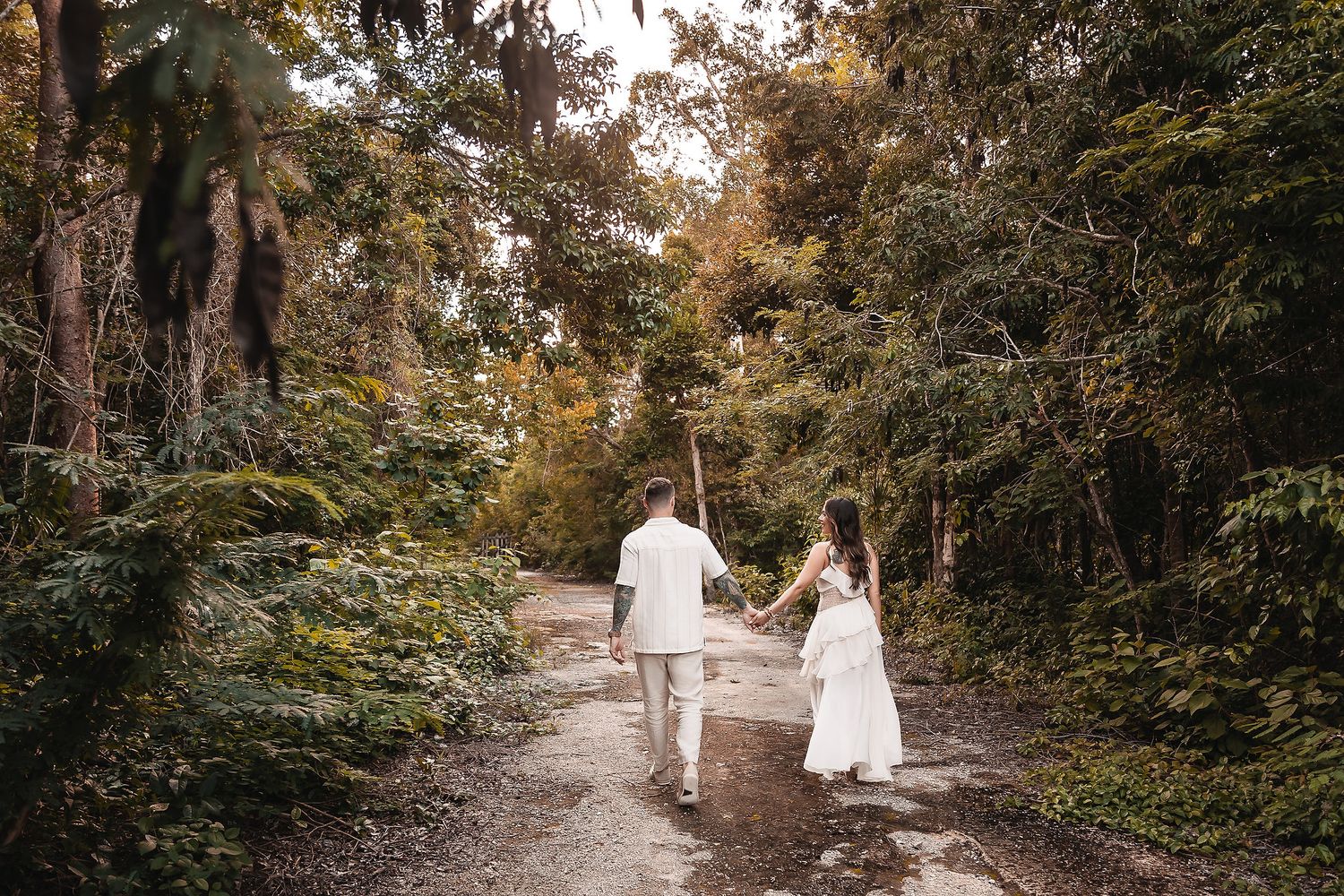 A couple in white outfits walks along a wooded path during an autumn photo session.