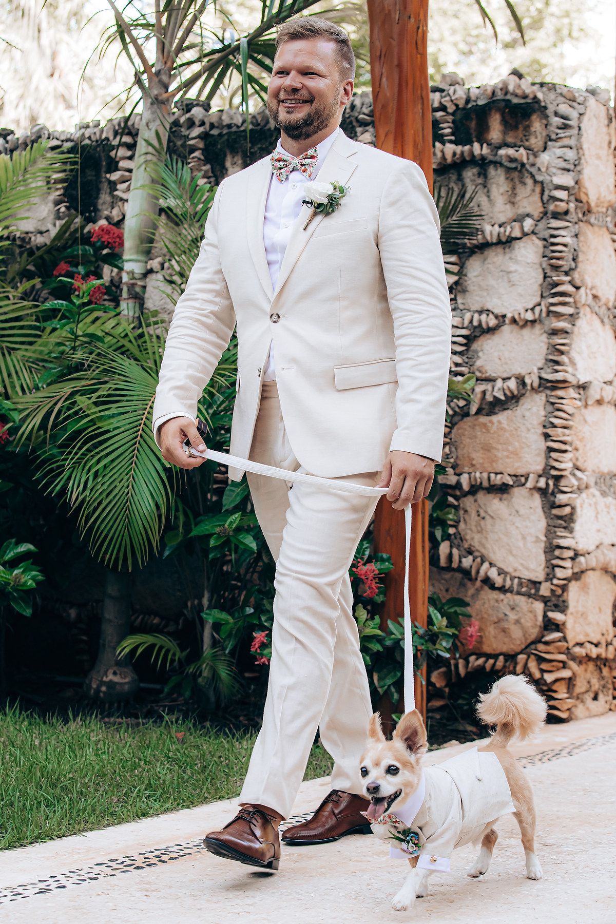 A small white dog walks alongside someone wearing a white suit and brown shoes on a garden path.