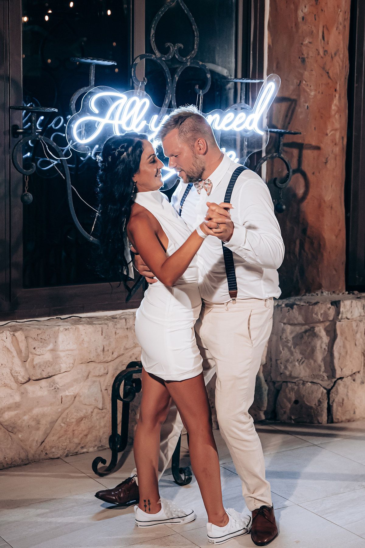 A couple in matching white outfits dances together against a brick wall with neon signage at night.
