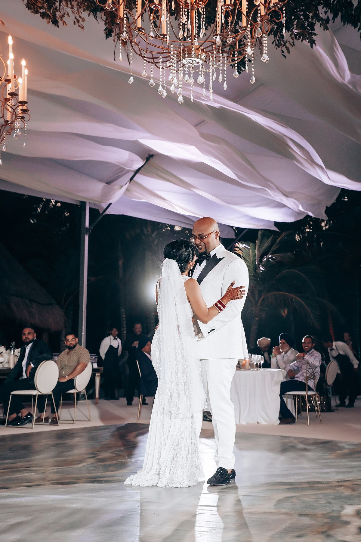 First dance at wedding reception under crystal chandeliers and flowing white fabric ceiling drapes.