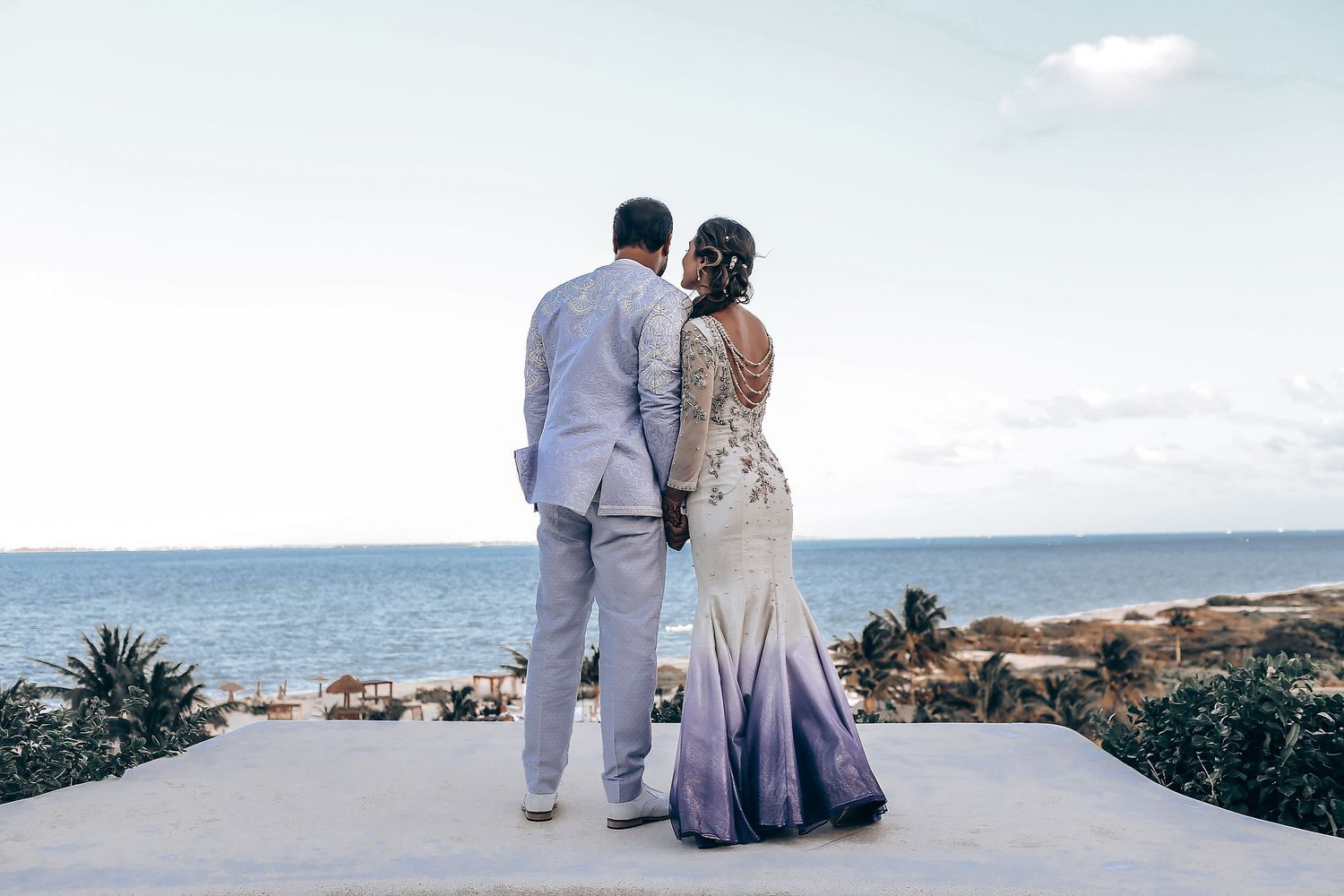 A couple in light-colored attire stands together looking out at the ocean on a sunny beach day.