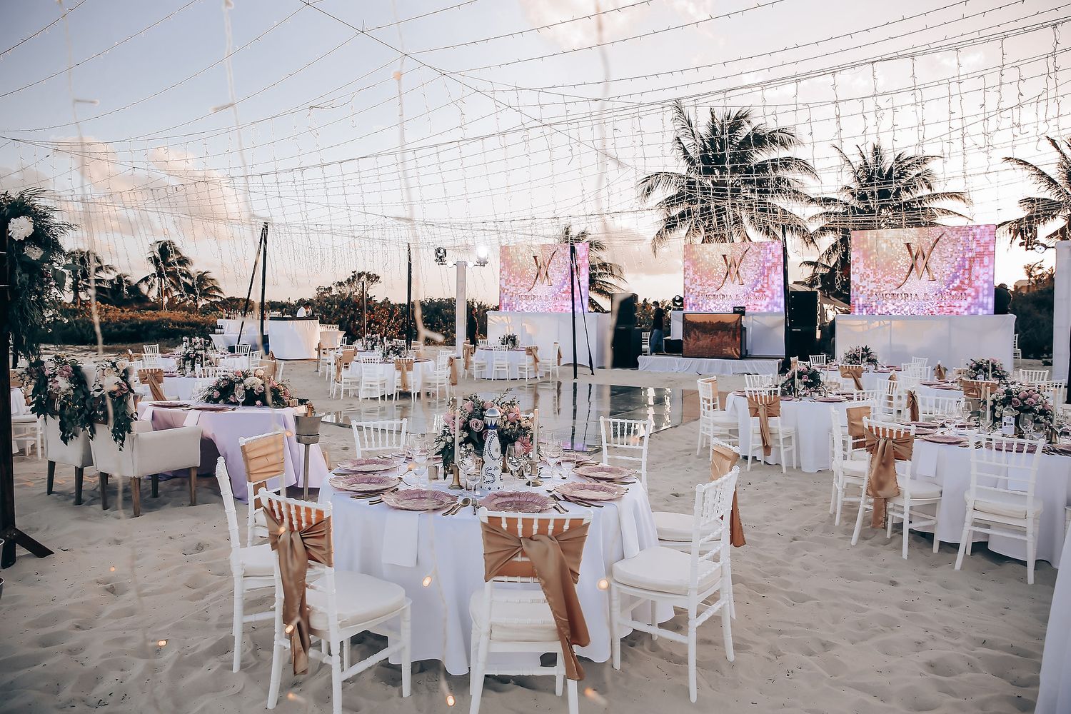 Elegant beach wedding reception setup with white chairs and tables decorated with palm trees and ocean views.