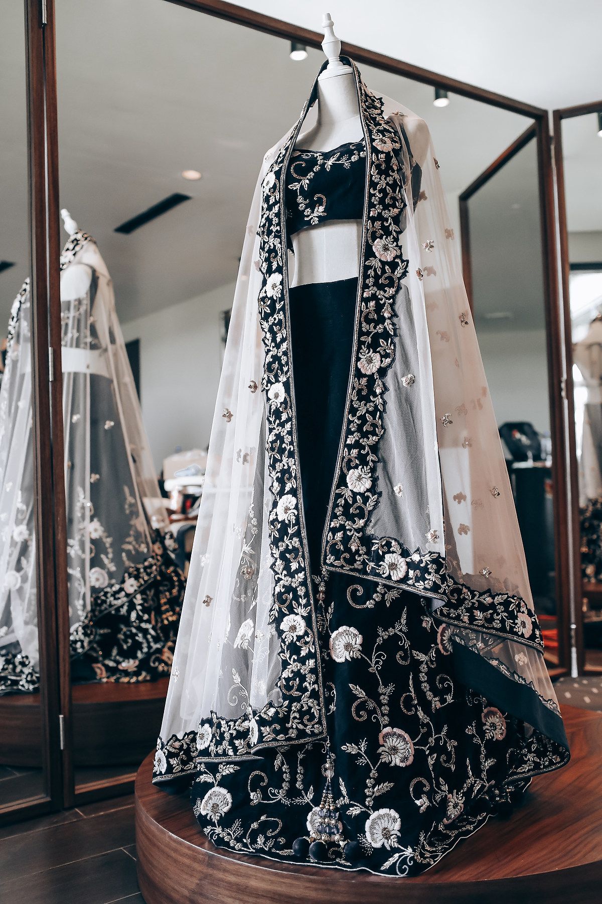 Black and white formal gown with intricate floral embroidery displayed on a wooden platform in a mirrored room.