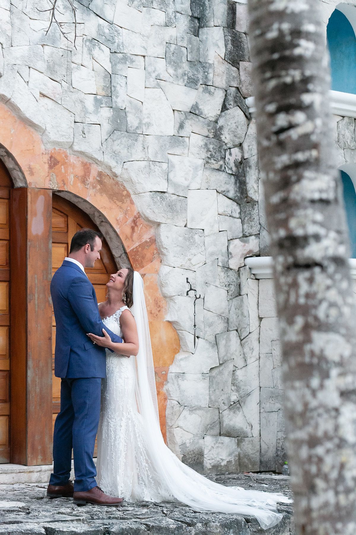 Wedding couple poses against rustic stone walls and wooden doors of a historic building.