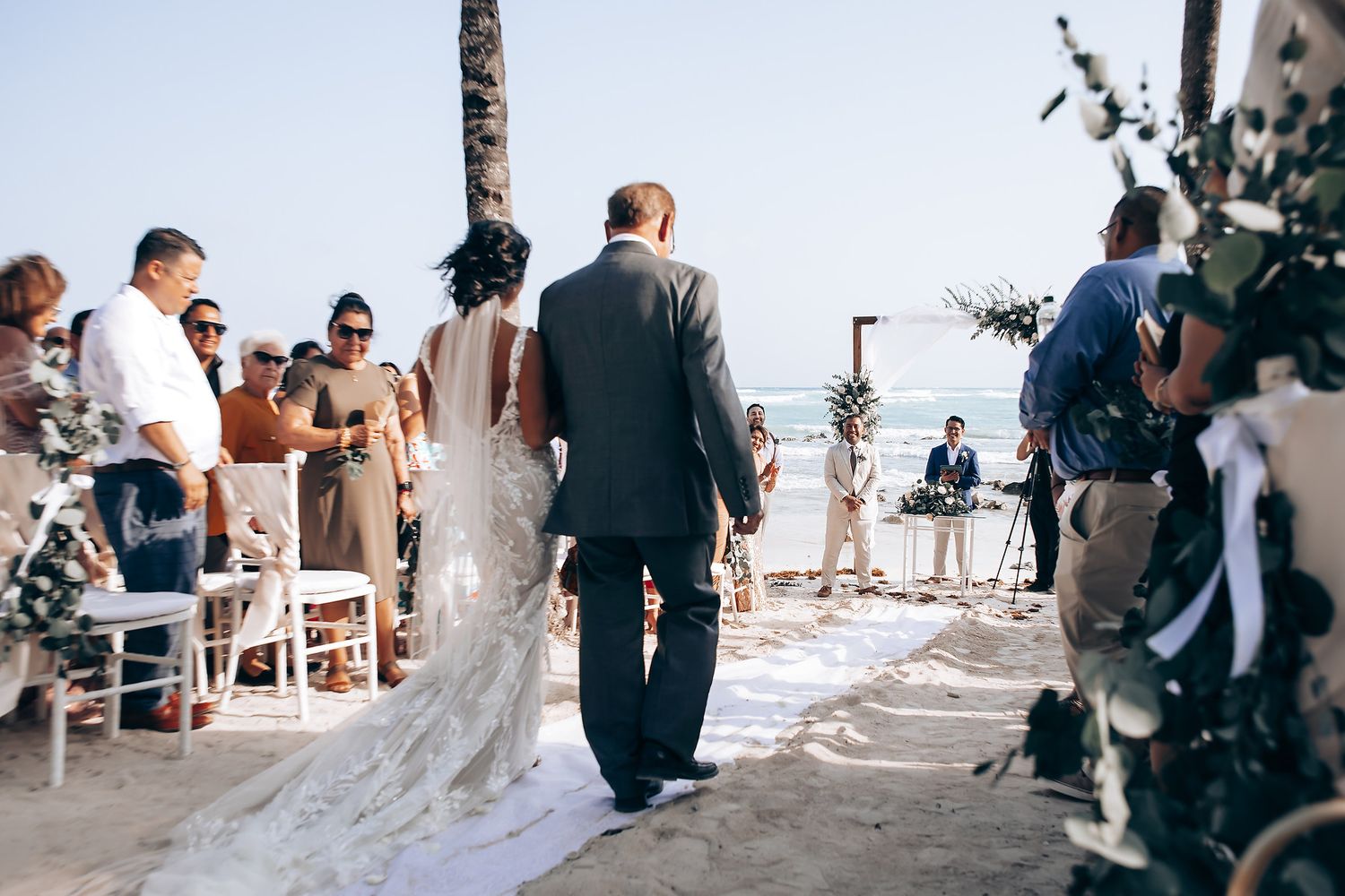 A wedding ceremony procession on a sandy beach with floral decorations and seated guests.