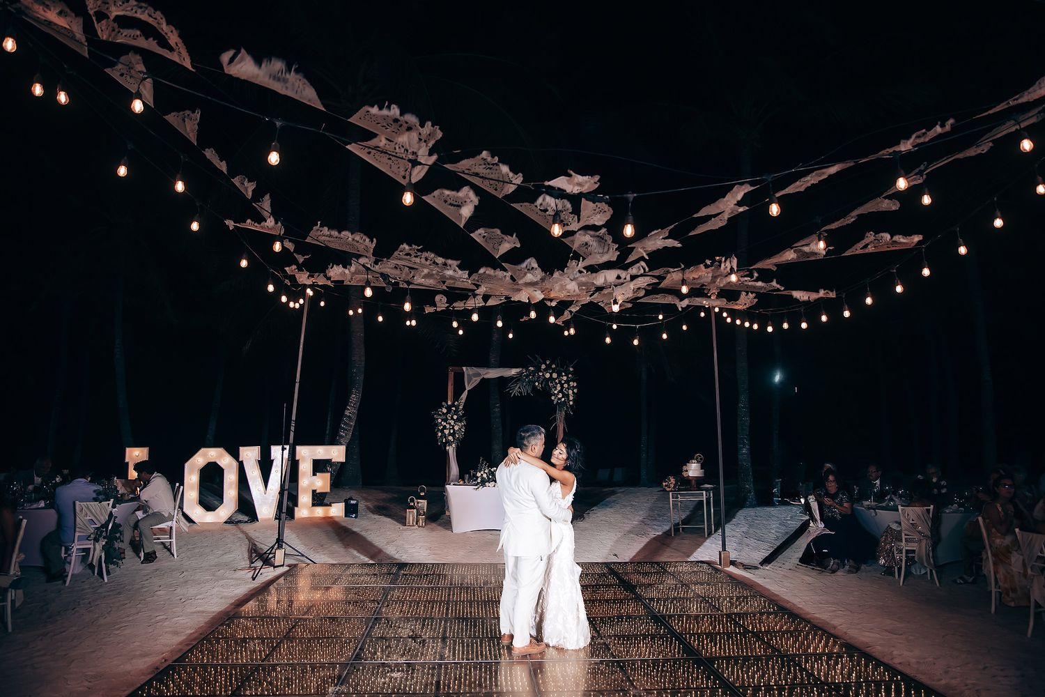 Romantic dance under illuminated driftwood ceiling with LOVE sign at beach wedding.