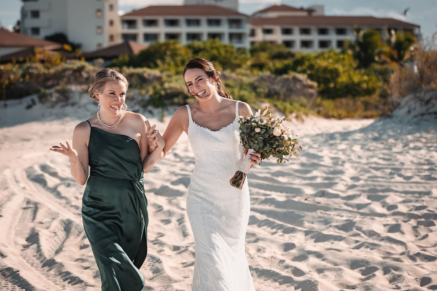 Two people walk together joyfully on a sandy beach with resort buildings in the background.