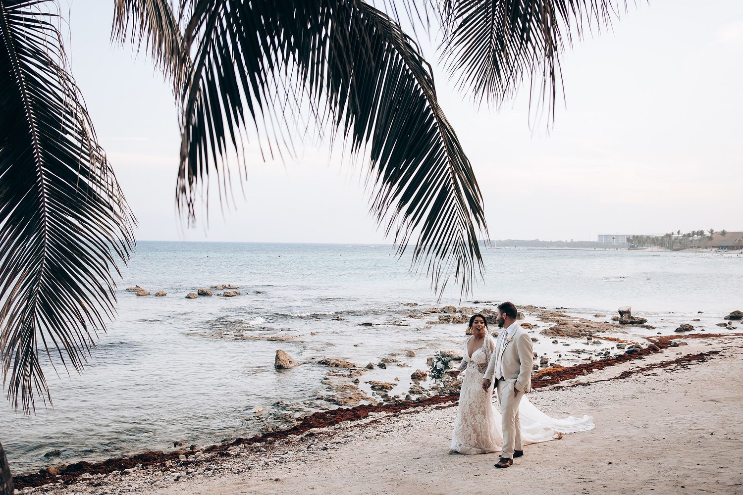 Couple walking along a tropical beach with palm trees and ocean views at sunset.