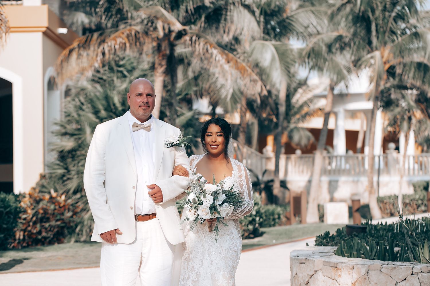 Couple celebrates their wedding day in a lush outdoor setting with palm trees and white decor.