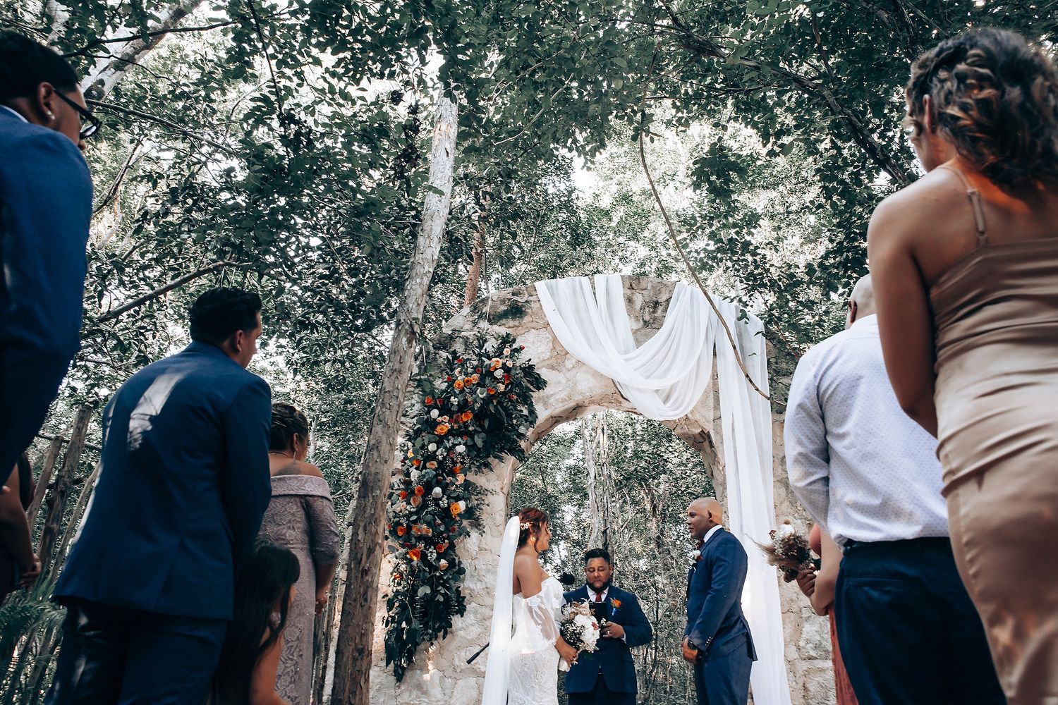Wedding guests gather in a natural forest setting with dappled sunlight filtering through trees.