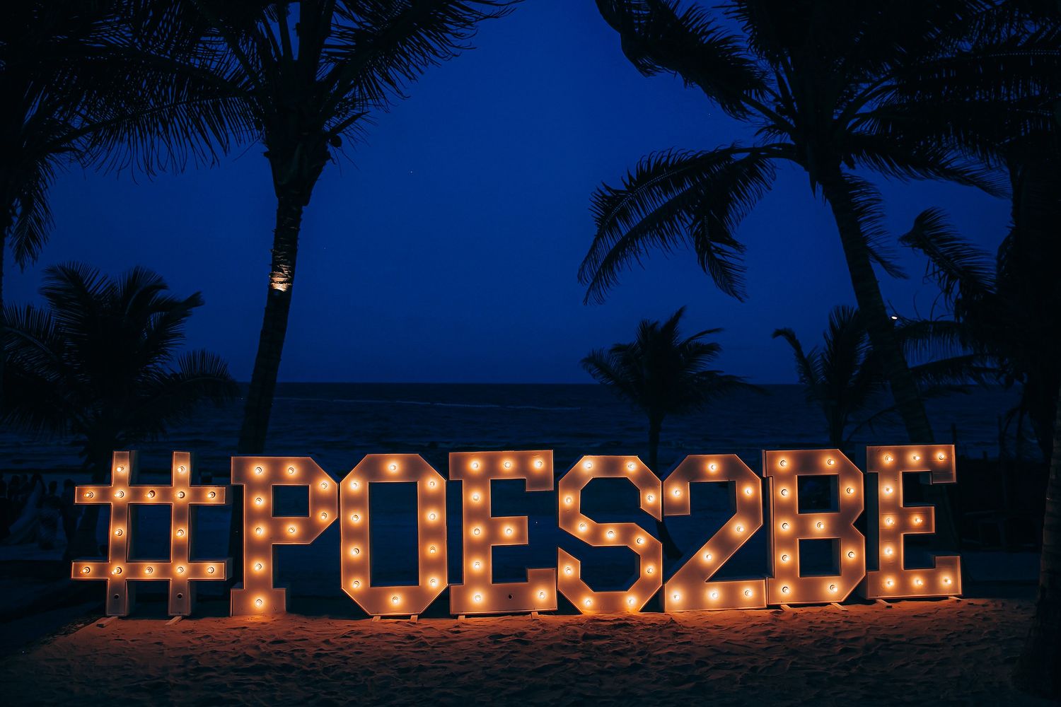 Illuminated letters spelling out a message with candles on a beach at night with palm trees silhouetted against a dark blue sky.