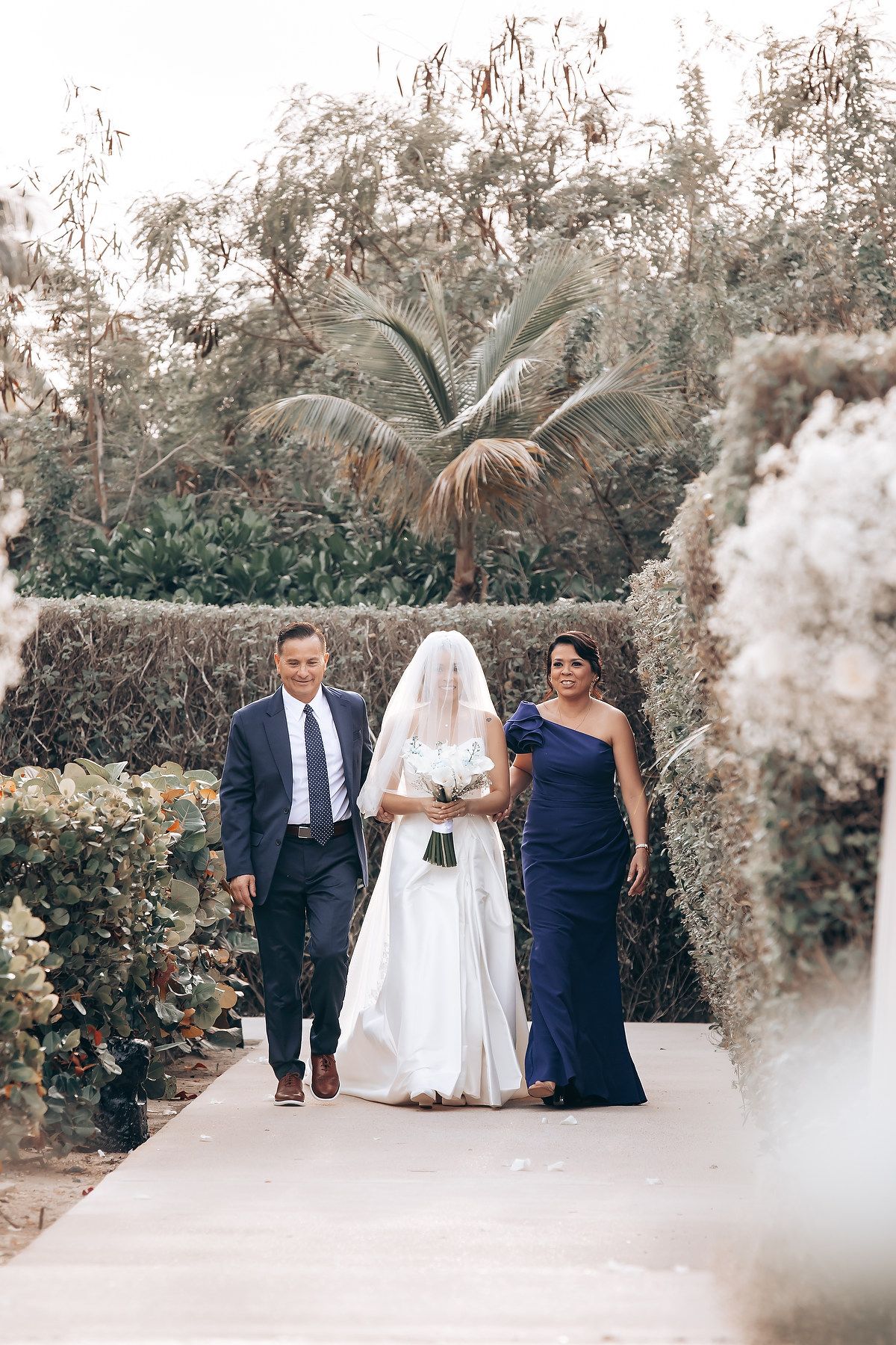 A wedding party walks down a tropical garden path lined with palm trees and white flowers.