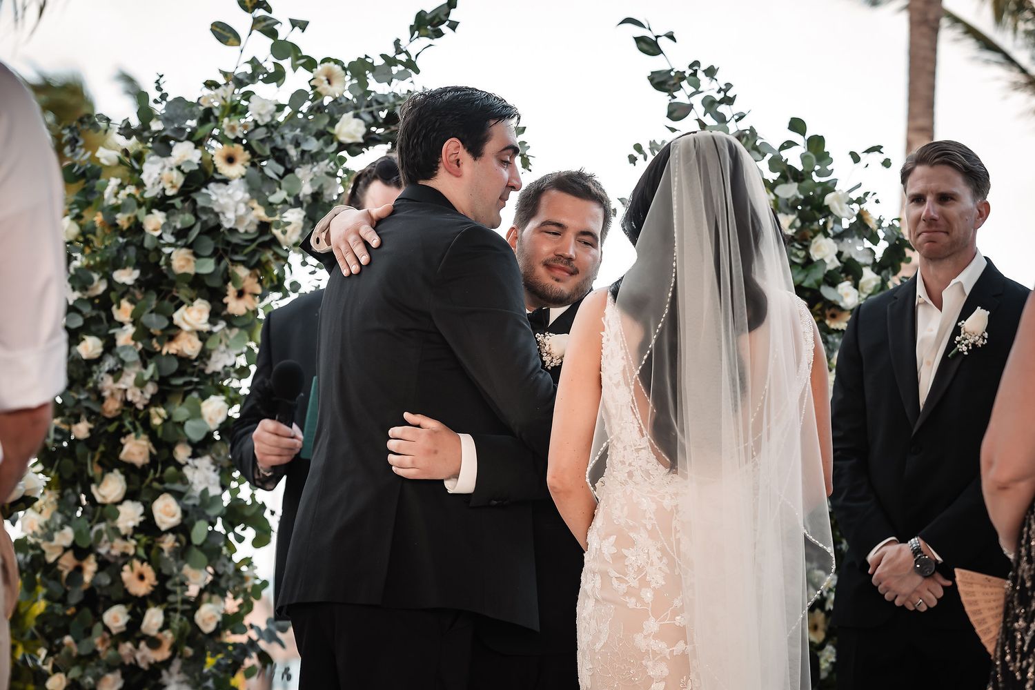A wedding ceremony setup with white floral arrangements and guests gathered outdoors.