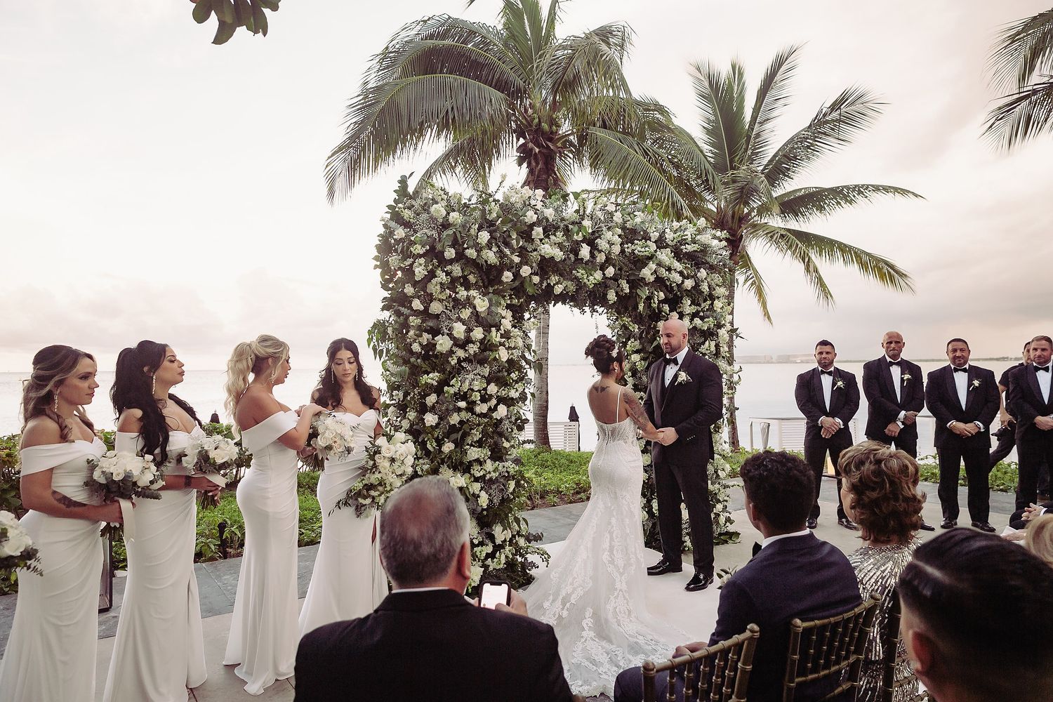 Wedding ceremony guests stand in a line under palm trees and floral arrangements during an outdoor celebration.