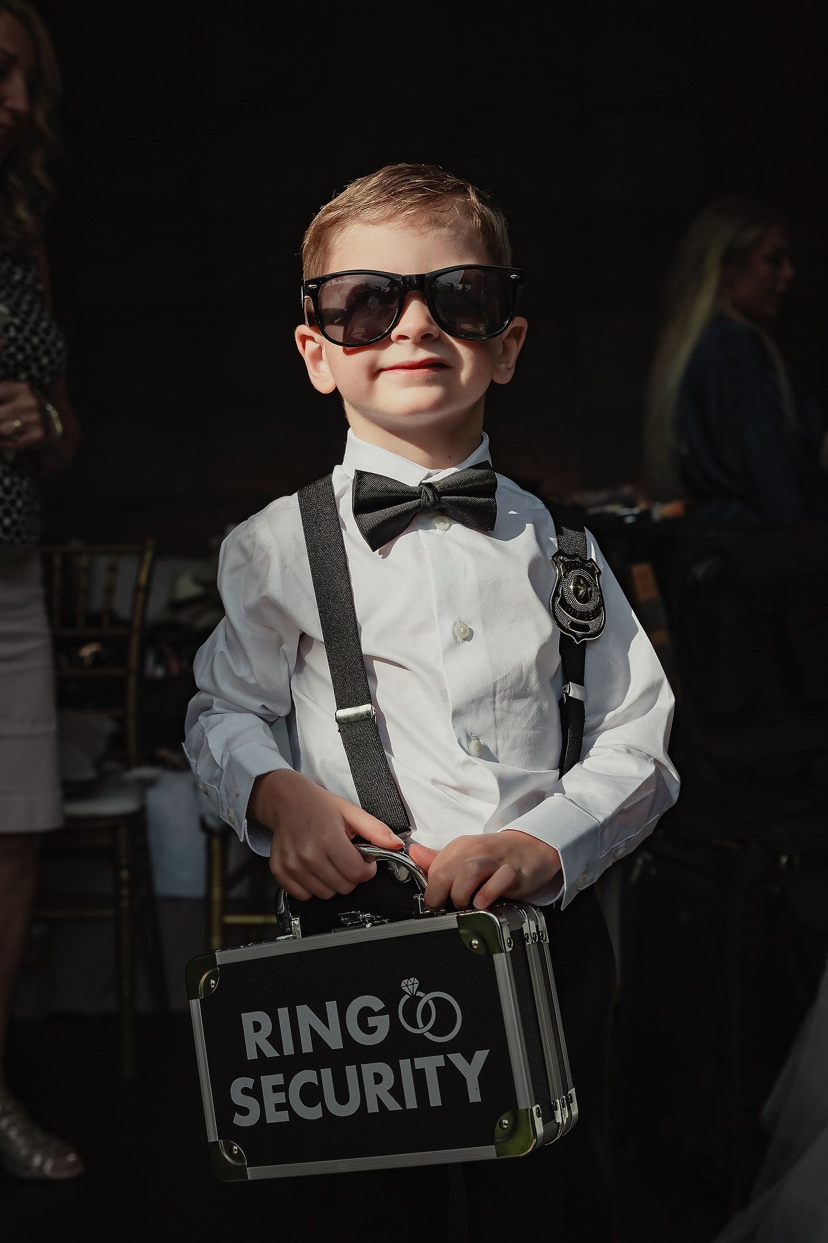 Young ring bearer in sunglasses, white shirt and suspenders holds security sign at wedding ceremony.