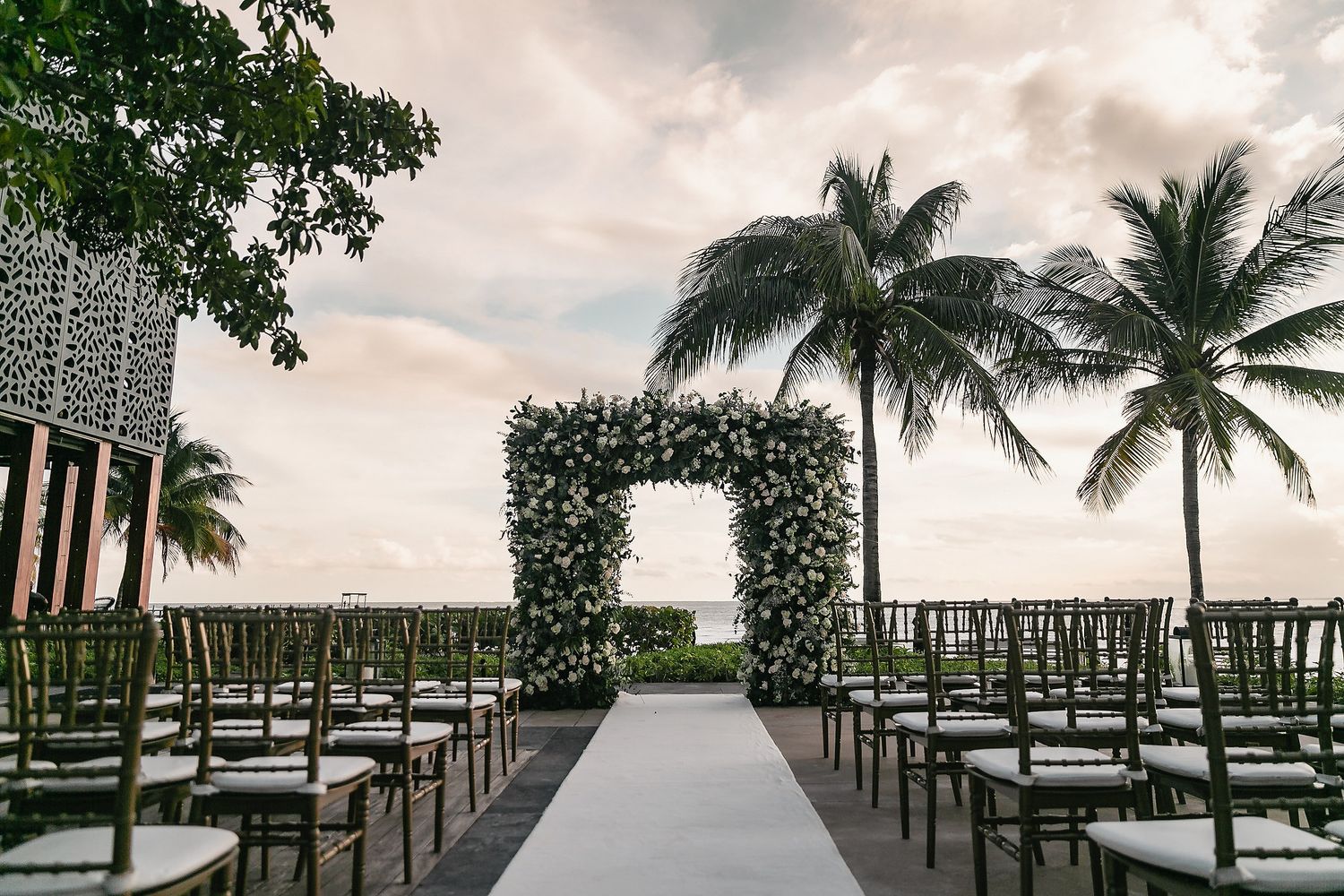 A scenic waterfront boardwalk with palm trees and boat docks stretches along the calm waters under cloudy skies.
