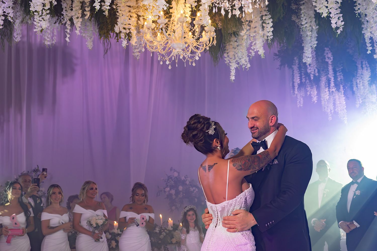 A wedding dance floor filled with guests celebrating under elegant crystal chandeliers and purple lighting.