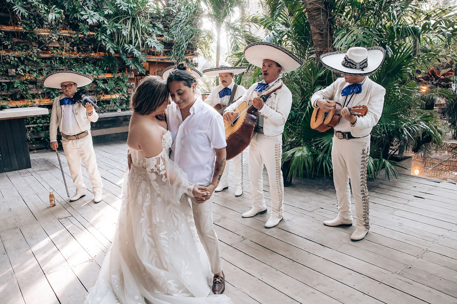 Wedding party in white attire dances on an outdoor terrace surrounded by tropical greenery.
