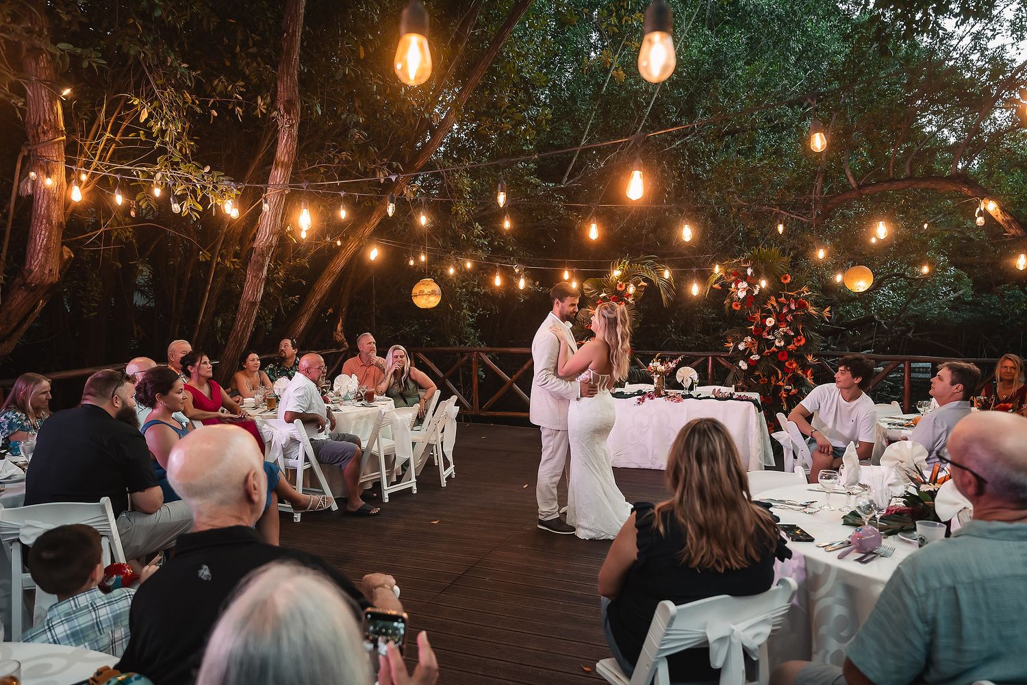 Newlyweds dancing on outdoor deck surrounded by guests and string lights.