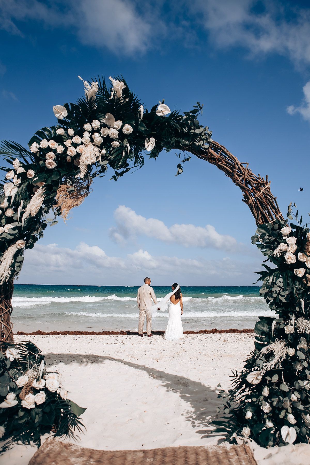 White floral wedding arch on sandy beach with turquoise ocean waves in background.