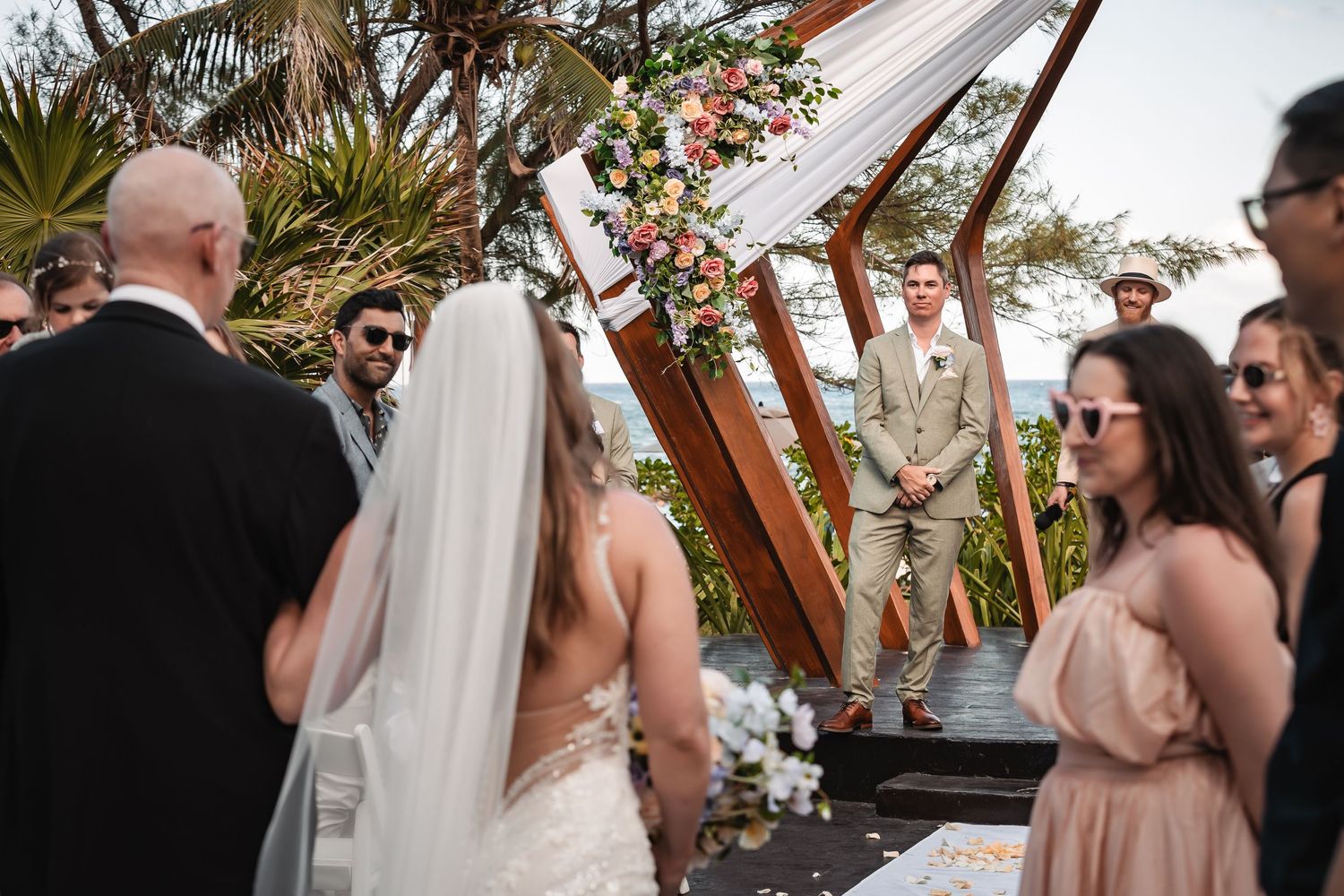 Outdoor wedding ceremony with guests seated under a modern wooden pergola.