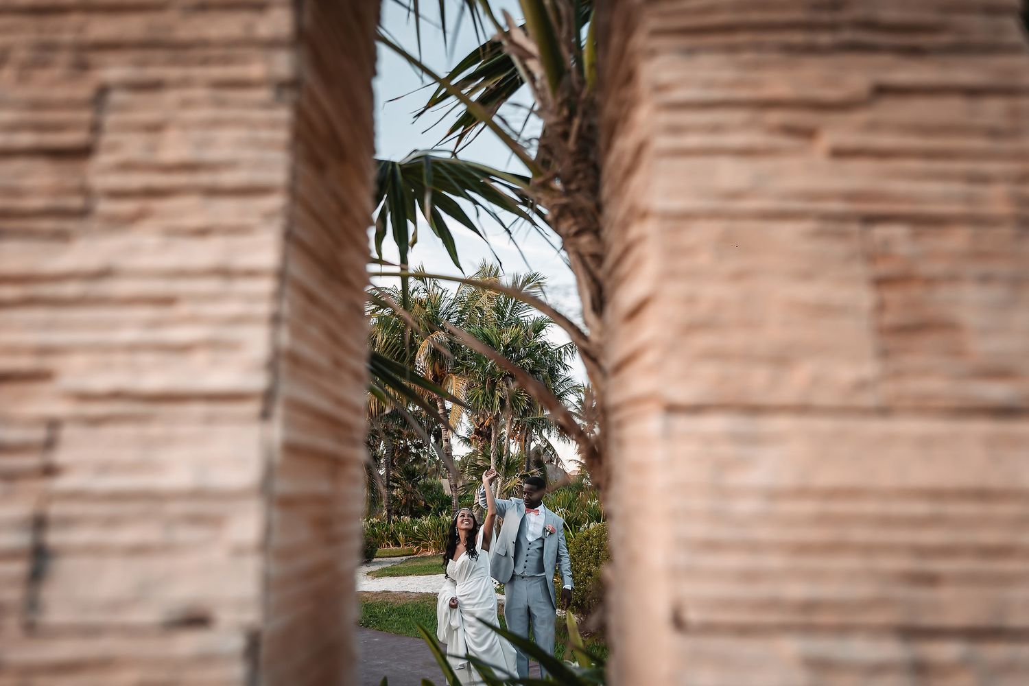 Wedding party glimpsed through stone pillars with palm trees in background.