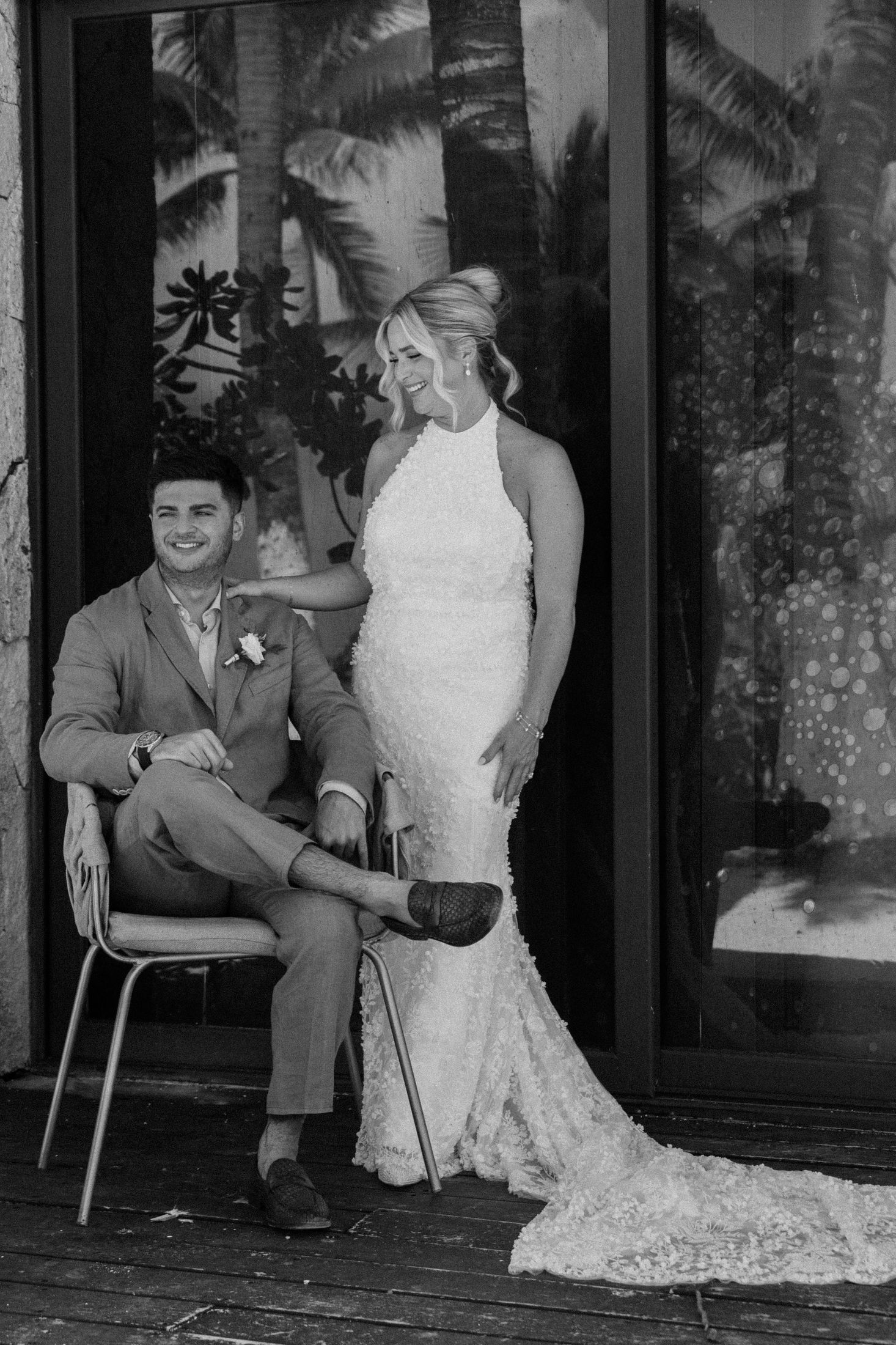 Black and white photo of bride in lace gown standing beside seated groom in suit at elegant wedding.