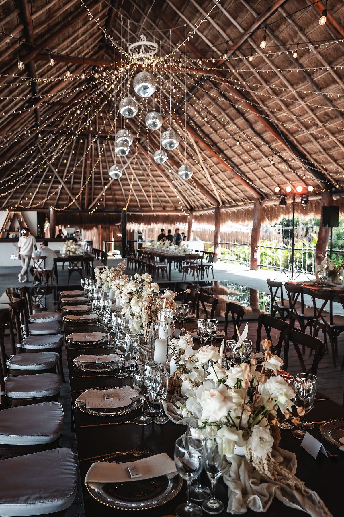Elegant wedding reception under a thatched roof with string lights, disco balls, and white floral centerpieces.