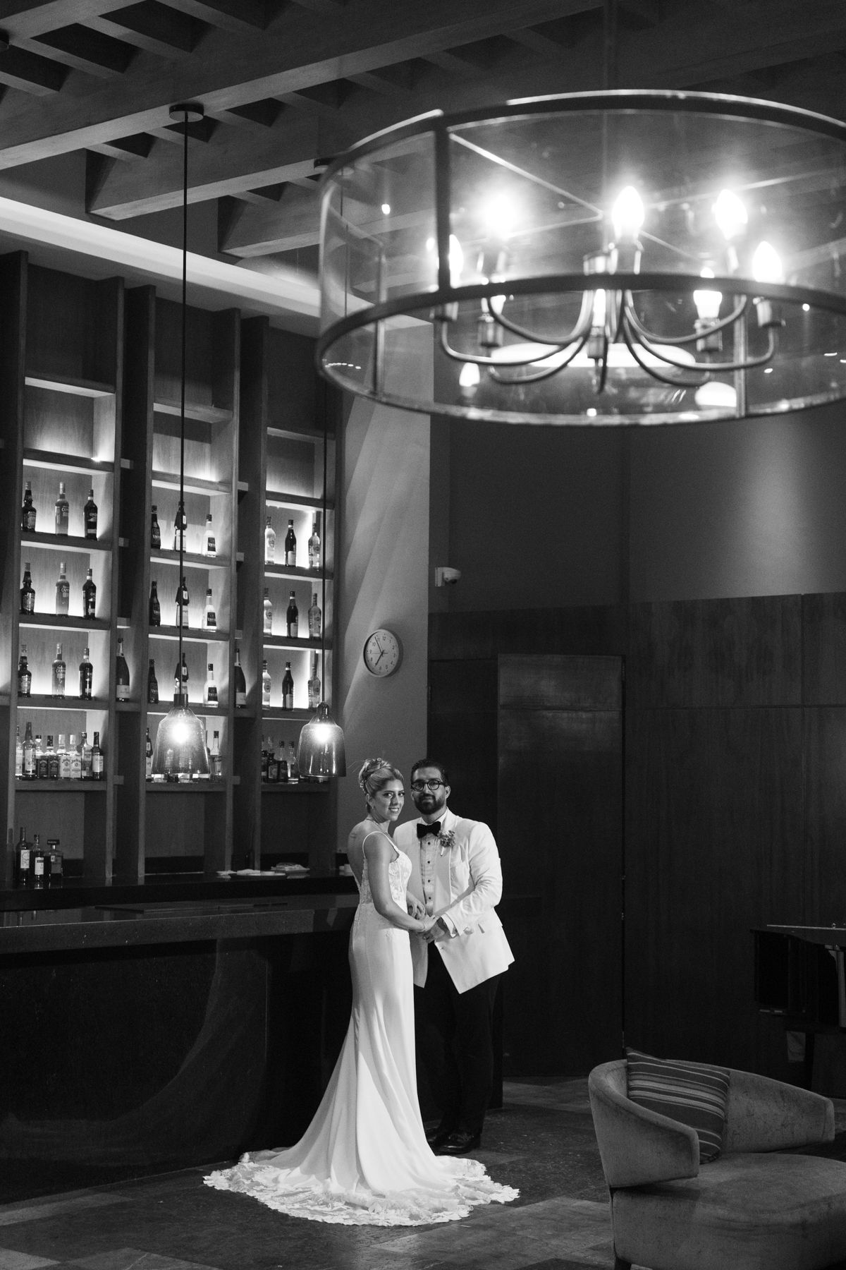 Black and white photo of bride and groom posing elegantly under chandelier in upscale bar venue.